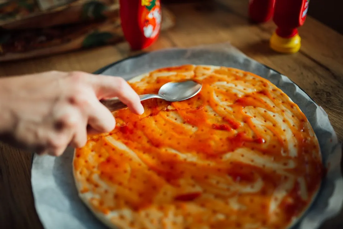 Hands spreading sauce on a homemade pizza